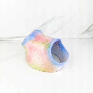 May include: A unique, handmade ceramic sculpture with a tunnel-like opening. The piece features a blend of pastel colors, including pink, blue, and green, creating a soft, artistic aesthetic. The sculpture's form is organic and textured.