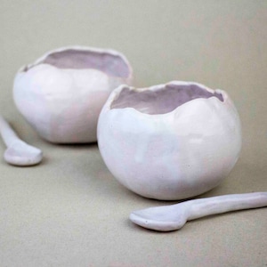 Puede incluir: Dos cuencos de cerámica blanca con una superficie ligeramente texturizada y dos cucharas de cerámica blanca a juego. Los cuencos están ligeramente deformados y tienen un aspecto rústico.