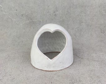 May include: A white ceramic object with a rounded, dome-like shape and a heart-shaped opening on the front. The surface has a textured, uneven finish with visible indentations, and the interior is hollow with a smooth, light-coloured base.