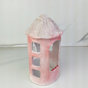 May include: A small, pink ceramic house with a textured, light-coloured roof. The house has three rectangular windows and a large opening on one side. The ceramic has a speckled appearance.