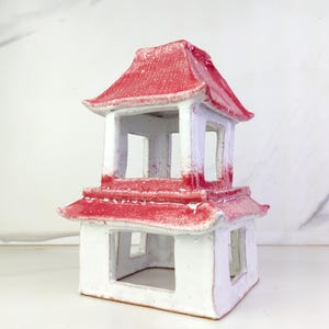 May include: A two-tiered ceramic pagoda with a red roof and white walls. The pagoda features open windows and a traditional Asian architectural design. The red roof has a textured appearance, and the white walls have a slightly speckled finish.