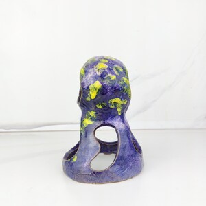 May include: A ceramic sculpture in the shape of a stylized figure. The sculpture is primarily a deep blue color, with splotches of yellow and green on the top. It has two large, open cutouts on the sides.