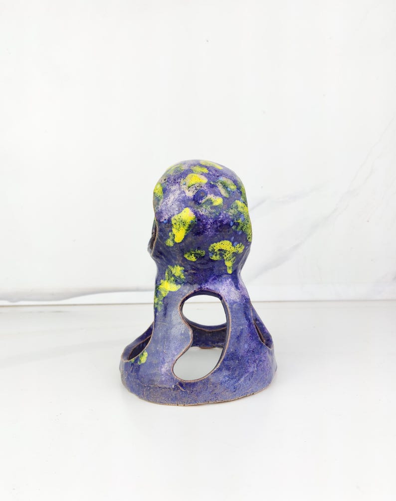 May include: A ceramic sculpture in the shape of a stylized figure. The sculpture is primarily a deep blue color, with splotches of yellow and green on the top. It has two large, open cutouts on the sides.