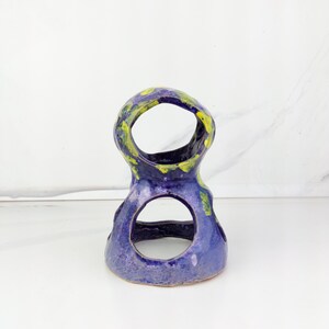 May include: A handmade ceramic sculpture in a unique shape, featuring a deep blue base with yellow and green accents. The sculpture has two large circular openings and a textured surface, perfect for home decor.