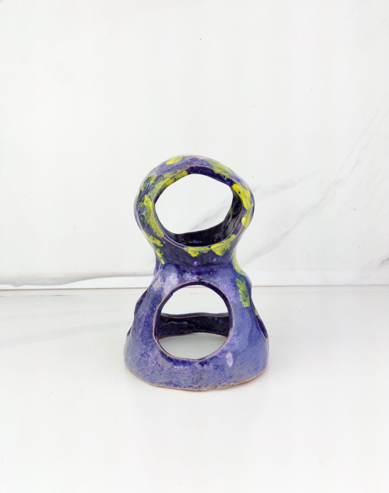 May include: A handmade ceramic sculpture in a unique shape, featuring a deep blue base with yellow and green accents. The sculpture has two large circular openings and a textured surface, perfect for home decor.