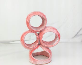 May include: A ceramic sculpture featuring four interconnected circular openings arranged in a vertical cluster. The object has a textured, mottled pink and white glaze with a glossy finish, creating a distinct, organic, and irregular shape.