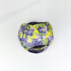 May include: A handmade ceramic vessel with a unique, organic shape. The exterior features a mottled pattern of yellow, purple, and blue hues. The vessel sits on a matching base, creating a cohesive and artistic piece. This decorative item is suitable for home decor.