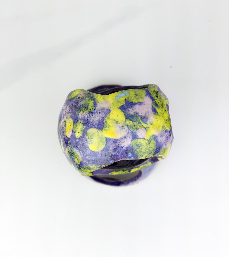 May include: A handmade ceramic vessel with a unique, organic shape. The exterior features a mottled pattern of yellow, purple, and blue hues. The vessel sits on a matching base, creating a cohesive and artistic piece. This decorative item is suitable for home decor.