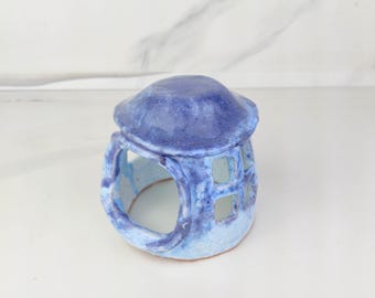 May include: A small, hand-sculpted ceramic structure featuring a rounded, dark blue roof and a lighter blue base. The piece has a wide, open arched entrance and a side wall with two square cut-out windows, showcasing a glossy, mottled glaze finish.