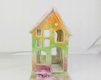 May include: A ceramic house-shaped candle holder featuring a speckled finish in shades of orange, green, and purple. The structure includes a gabled roof, multiple rectangular window cutouts, an arched doorway, and a small attached table.