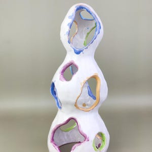 May include: A white ceramic sculpture with a stacked, organic form. The sculpture has several irregularly shaped openings, each outlined with colourful paint in shades of blue, purple, orange, and green. The piece has a textured surface.