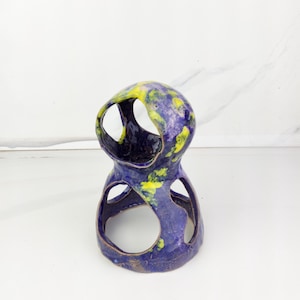 May include: A handcrafted ceramic sculpture with a unique, open design. The piece features a deep blue base with yellow and green accents, creating a marbled effect. The sculpture has multiple circular openings and a glossy finish, suitable for home decor.