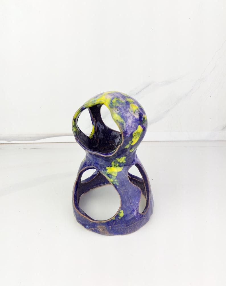 May include: A handcrafted ceramic sculpture with a unique, open design. The piece features a deep blue base with yellow and green accents, creating a marbled effect. The sculpture has multiple circular openings and a glossy finish, suitable for home decor.