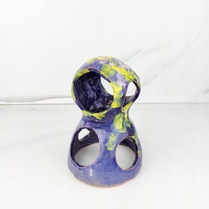 May include: A handcrafted ceramic sculpture with a unique, open design. The piece features a two-tiered structure with circular openings and is painted in shades of blue and yellow. The abstract form is suitable for home decor.
