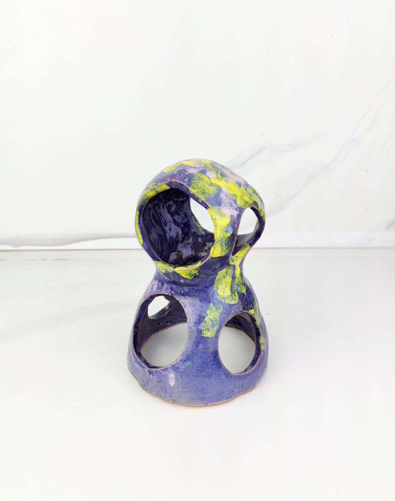 May include: A handcrafted ceramic sculpture with a unique, open design. The piece features a two-tiered structure with circular openings and is painted in shades of blue and yellow. The abstract form is suitable for home decor.
