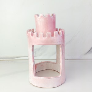 May include: A pink ceramic castle-shaped candle holder with a rectangular opening and a cylindrical tower. The castle has a textured surface and a white interior. The candle holder is on a white surface.