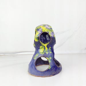 May include: A handcrafted ceramic sculpture in shades of blue, yellow, and purple. The unique piece features a rounded top with a series of circular openings. The sculpture's surface has a textured appearance, adding to its artistic appeal.