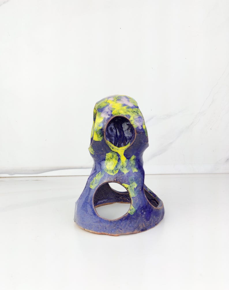 May include: A handcrafted ceramic sculpture in shades of blue, yellow, and purple. The unique piece features a rounded top with a series of circular openings. The sculpture's surface has a textured appearance, adding to its artistic appeal.