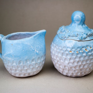 Pode incluir: Um conjunto de jarro de creme e açucareiro de cerâmica azul e branco. O jarro de creme tem uma alça e uma forma arredondada. O açucareiro tem uma tampa com uma pega arredondada. Ambas as peças têm uma superfície texturizada.