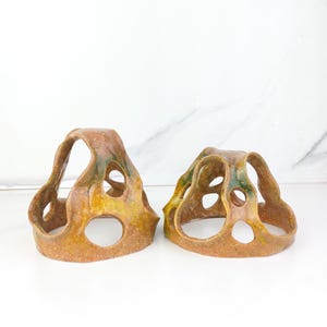 May include: Two hand-formed ceramic sculptures with an organic, hollow structure featuring multiple circular cutouts. The clay has a speckled, glazed finish in shades of earthy brown, ochre, and hints of deep green, with a smooth, glossy texture.