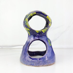 May include: A handmade ceramic sculpture in a figure-eight shape. The piece is primarily a deep blue with accents of yellow and green. The sculpture has two large circular openings and a textured surface.