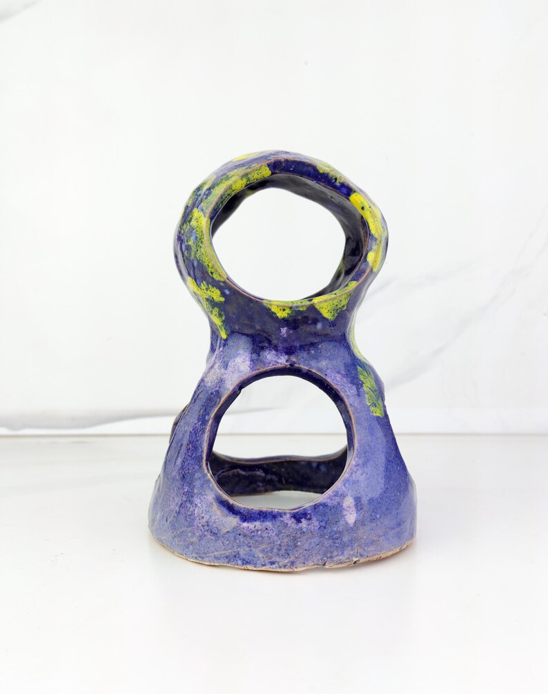 May include: A handmade ceramic sculpture in a figure-eight shape. The piece is primarily a deep blue with accents of yellow and green. The sculpture has two large circular openings and a textured surface.