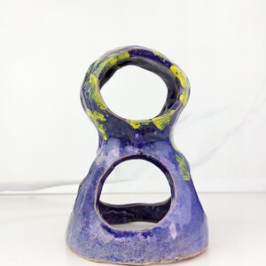 May include: A handmade ceramic sculpture with a unique, open design. The piece features a vibrant blue glaze with accents of yellow and green. The sculpture has two large circular openings and a textured surface, showcasing the artist's craftsmanship.