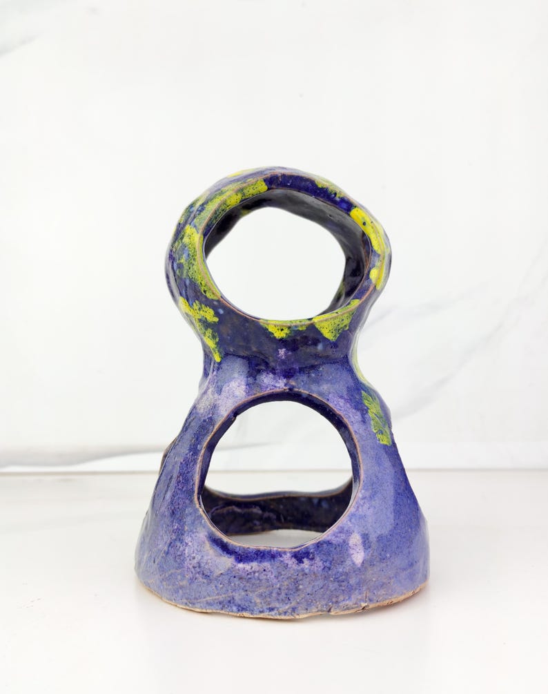 May include: A handmade ceramic sculpture with a unique, open design. The piece features a vibrant blue glaze with accents of yellow and green. The sculpture has two large circular openings and a textured surface, showcasing the artist's craftsmanship.