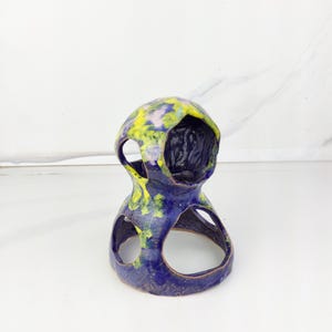 May include: A handcrafted ceramic sculpture in a unique, abstract design. The piece features a deep blue base with yellow and green accents on the top. It has two open circular cutouts and a central opening, showcasing a textured interior.