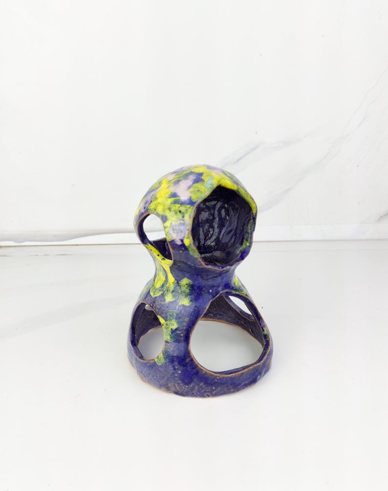 May include: A handcrafted ceramic sculpture in a unique, abstract design. The piece features a deep blue base with yellow and green accents on the top. It has two open circular cutouts and a central opening, showcasing a textured interior.