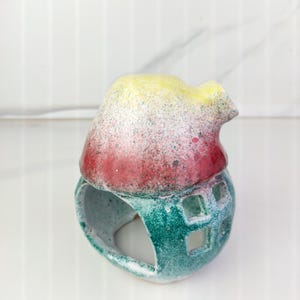 K&ouml;nnte beinhalten: Ein keramikhausf&ouml;rmiger Kerzenhalter. Das Dach ist gelb und rot mit gesprenkelter Oberfl&auml;che. Der Sockel ist gr&uuml;n mit quadratischen Aussparungen. Der Kerzenhalter ist f&uuml;r ein Teelicht konzipiert.