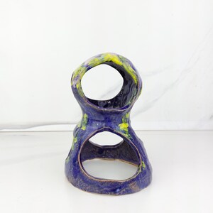 May include: A handmade ceramic sculpture in a unique, organic shape. The piece features a deep blue glaze with accents of yellow and green. It has two large openings, creating an open, airy design. The sculpture is placed on a white surface.