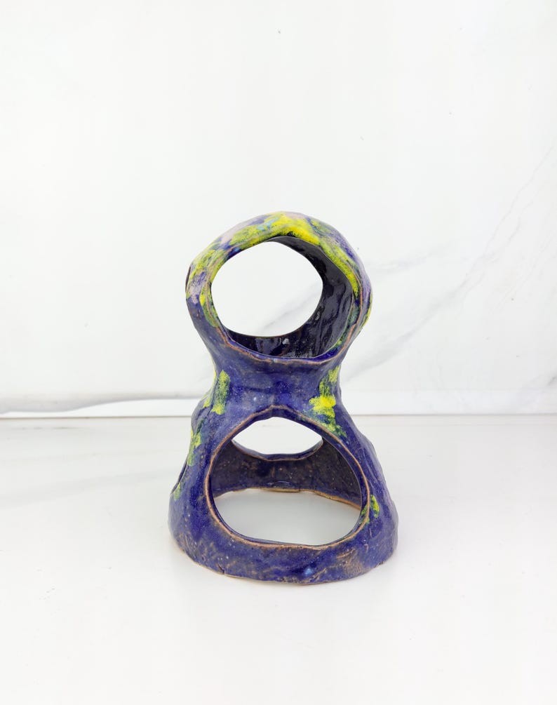 May include: A handmade ceramic sculpture in a unique, organic shape. The piece features a deep blue glaze with accents of yellow and green. It has two large openings, creating an open, airy design. The sculpture is placed on a white surface.
