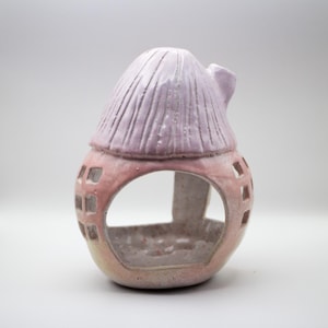May include: A ceramic house-shaped candle holder with a pink and white glaze. The house has a small, pointed roof and a cutout window.