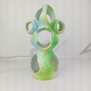 May include: A ceramic sculpture with a green and blue ombre glaze. The sculpture has multiple holes and a unique, abstract design.