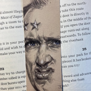 May include: A bookmark with a pencil sketch of a man's face, showing a serious expression and blue eyes. The bookmark has three star tattoos and a nose ring. It rests on an open book, with text visible on both sides.