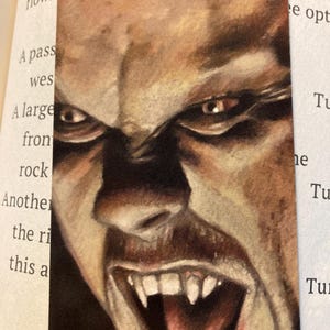 May include: A bookmark featuring a detailed, close-up illustration of a vampire's face. The artwork showcases a snarling expression with sharp fangs, intense eyes, and a dramatic, dark color palette. The bookmark is designed for book lovers.