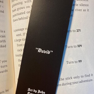 May include: A black bookmark with the name "David" in white gothic font. The bottom of the bookmark reads "Art by John Townshend" in a similar font. The bookmark is resting on the pages of a book.