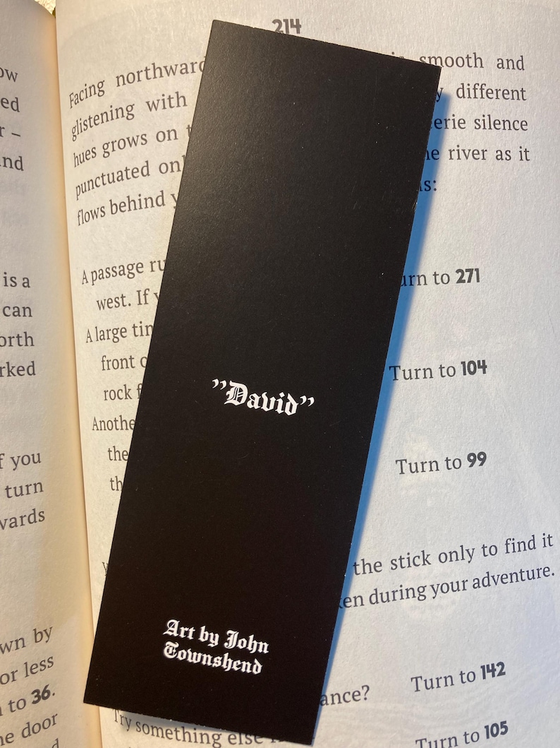 May include: A black bookmark with the name "David" in white gothic font. The bottom of the bookmark reads "Art by John Townshend" in a similar font. The bookmark is resting on the pages of a book.