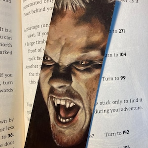 May include: A bookmark featuring a detailed illustration of a man's face with an angry expression, bared teeth, and sharp fangs. The artwork is set against a dark background, with the bookmark placed in an open book.