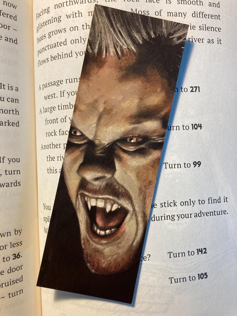 May include: A bookmark featuring a detailed illustration of a man's face with an angry expression, bared teeth, and sharp fangs. The artwork is set against a dark background, with the bookmark placed in an open book.