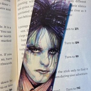 May include: A bookmark with a watercolour portrait of a person with dark hair and dramatic eye makeup. The image has a gothic aesthetic, with a sombre expression. The bookmark is placed on an open book.