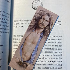 May include: A rectangular bookmark with an illustration of a man with long hair and a partially unbuttoned blue shirt. The bookmark is placed on an open book, with text visible on the pages.