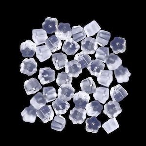 May include: A pile of clear, translucent earring backs, set against a black backdrop. The earring backs feature a flower-shaped design on one side and a square shape on the other, designed to securely hold earrings in place.