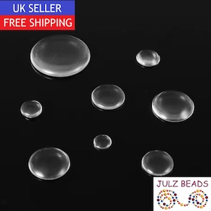 May include: A collection of seven clear, circular, dome-shaped cabochons of varying sizes arranged on a black surface. These smooth, transparent, glass-like pieces feature a rounded top and a flat base, reflecting light across their curved surfaces.