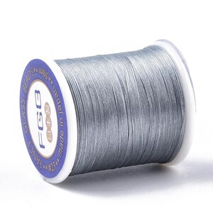 May include: A grey thread spool for crafts. The spool has a white plastic core with a blue label that reads "FANCY GLASS BEADS" and "made in Japan". The thread is tightly wound and appears smooth.