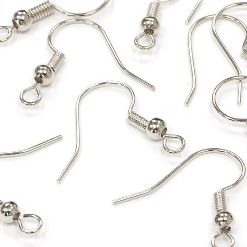 May include: Close-up of several silver-colored earring hooks, scattered against a white background. Each hook features a coiled spring detail and a small, round bead. The hooks are designed for jewelry making and crafting.