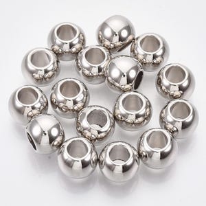 May include: A collection of shiny, silver-coloured metal beads. Each bead is spherical with a large, central hole and decorative detailing around the edges. The beads are reflective, creating highlights and shadows.