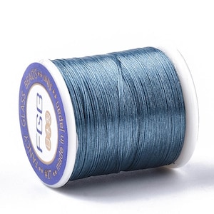 May include: A spool of blue-grey thread for crafting. The label reads "FGB" and "made in Japan". The thread is tightly wound on a white plastic spool. The text "Fancy Glass Beads" is also visible on the label.