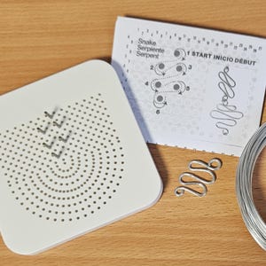 May include: A white beading board with a spiral pattern and directional arrows, a silver wire snake-shaped bead, a diagram with the text "Snake Serpentine Serpent" and a coil of silver wire. Craft supplies.
